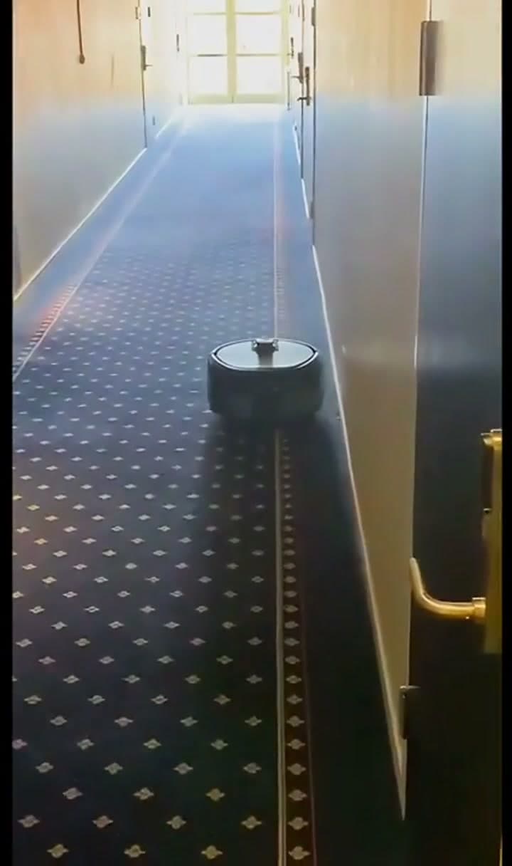 Kompakt rengjøringsrobot arbeider langs kanten av en hotellkorridor.