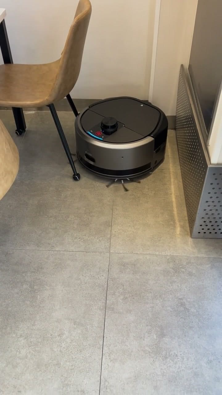 Kompakt rengjøringsrobot arbeider på gulvet mellom bord og fast inventar i et serveringslokale.