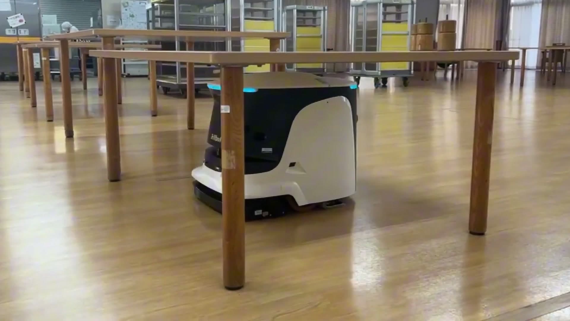 En kompakt rengjøringsrobot arbeider mellom bord i et tomt fellesareal på et sykehjem.