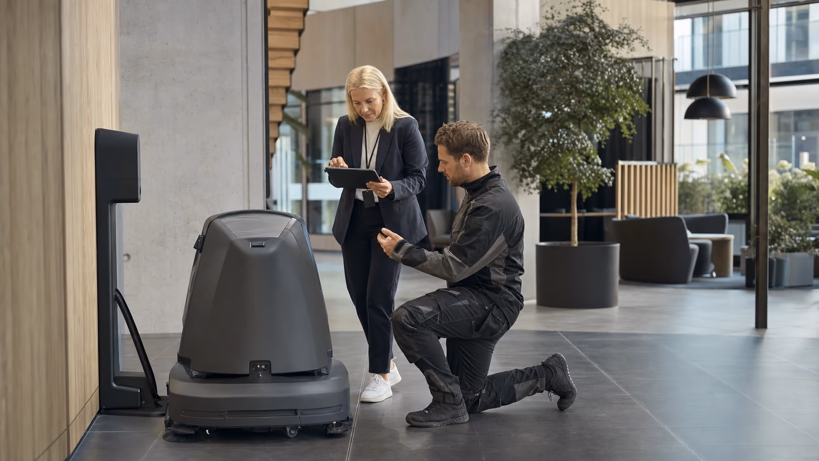 En driftsleder og en servicetekniker følger opp en profesjonell rengjøringsrobot ved ladestasjonen.